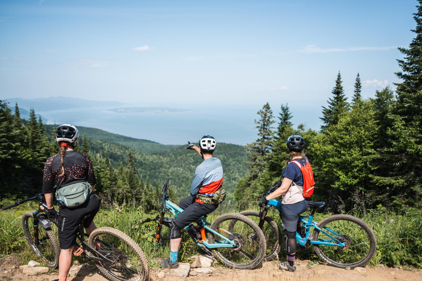 Trois cyclistes admirent une vue montagneuse, forêt et mer au loin.