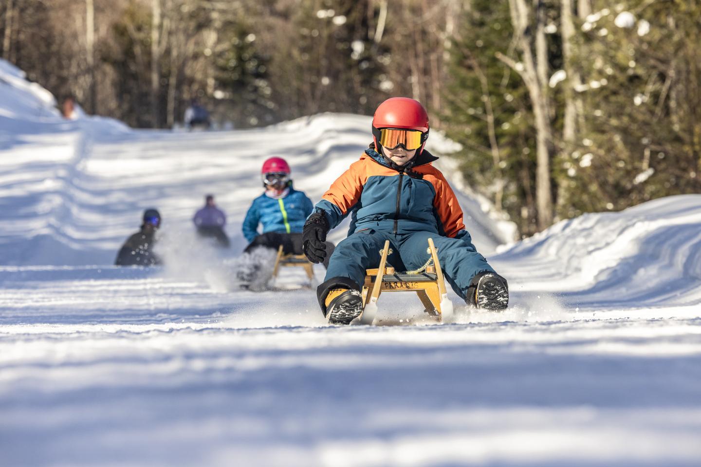 Enfants en luge sur une piste enneigée, forêt en arrière-plan.