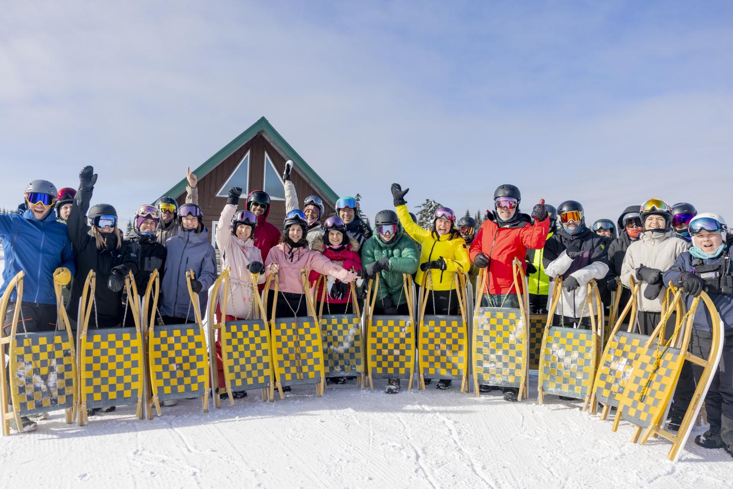 Groupe de personnes en tenue de ski, souriant avec des luges, ciel dégagé en arrière-plan.