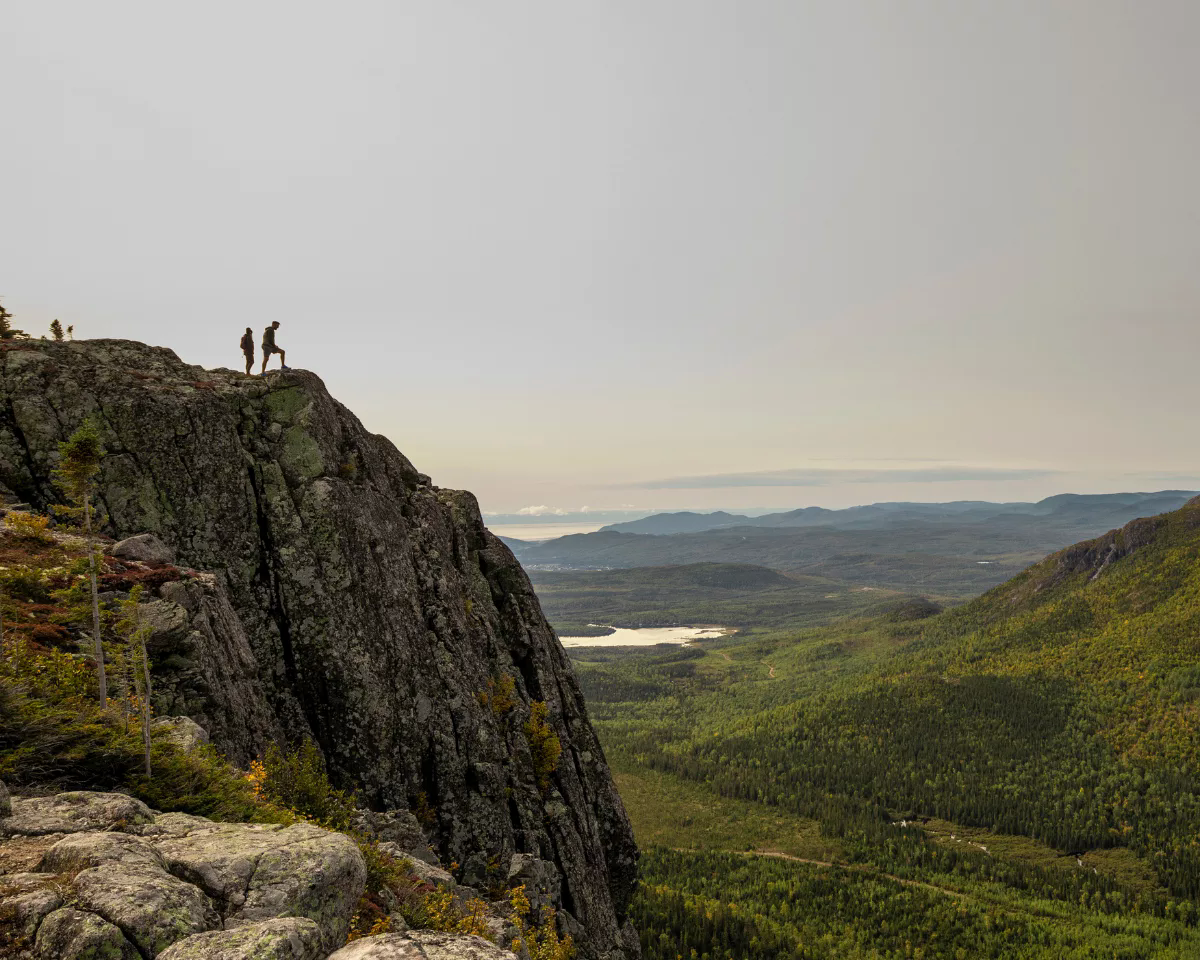 Deux randonneurs sur une falaise, vue panoramique sur une vallée verdoyante.